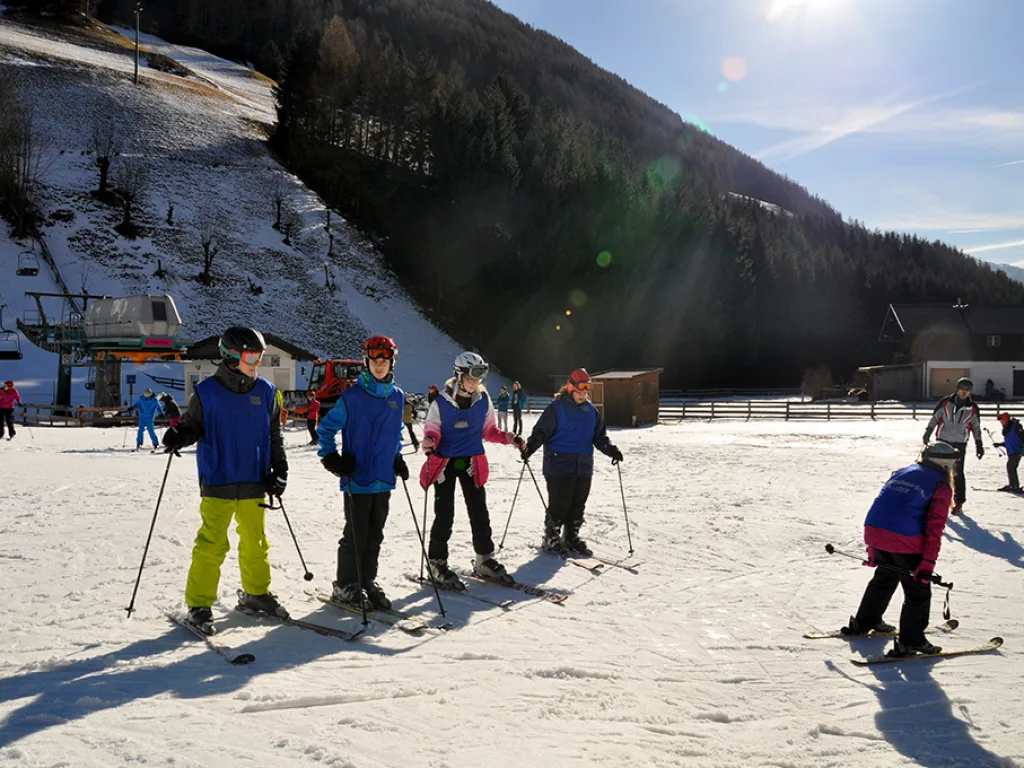Skigruppe steht am Pistenrand vor einem bewaldeten Hang in der Morgensonne.